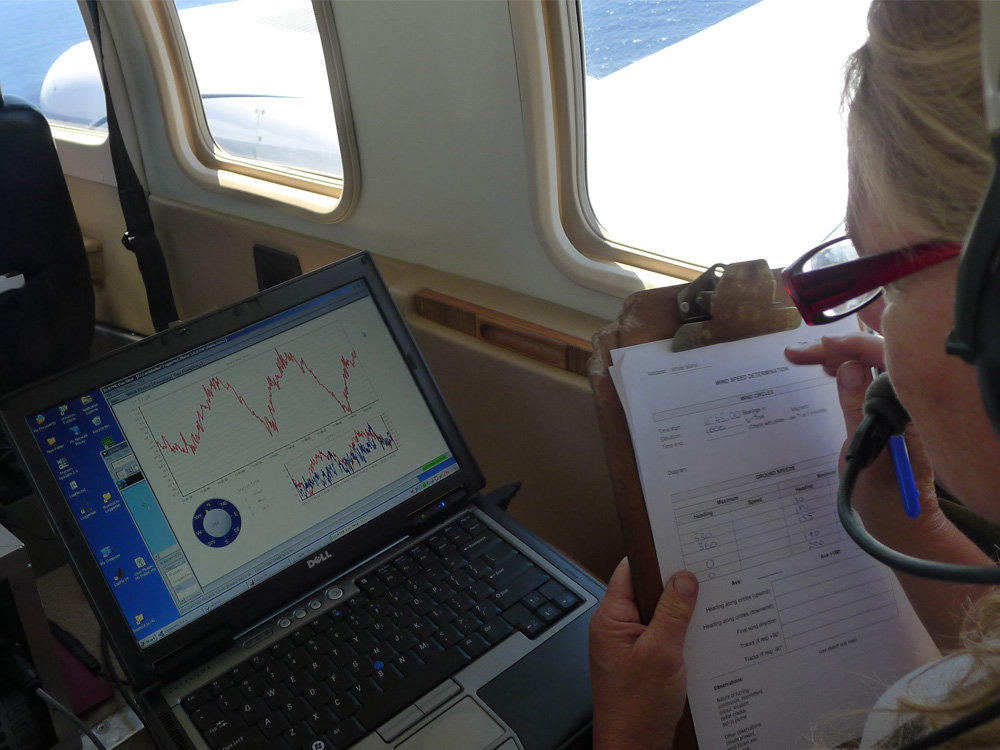 A volcanologist in a plane monitors their gas collection system using a computer display from the collection system. They have a clipboard and are noting relevant information.