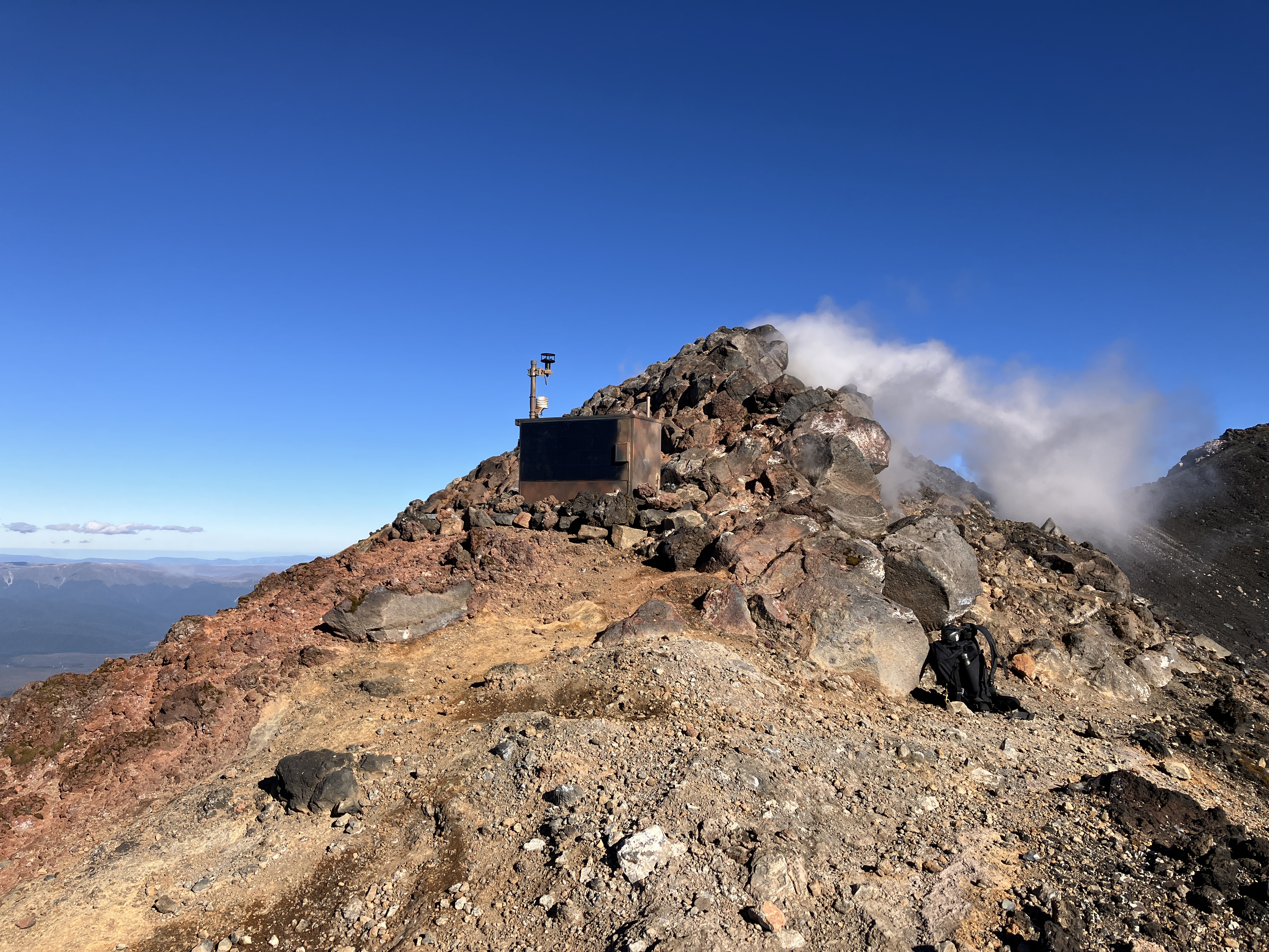 A cabinet containing a multigas system highlighted on the skyline close to the summit of Ngauruhoe, with steam from a fumarole drifting away in the wind.