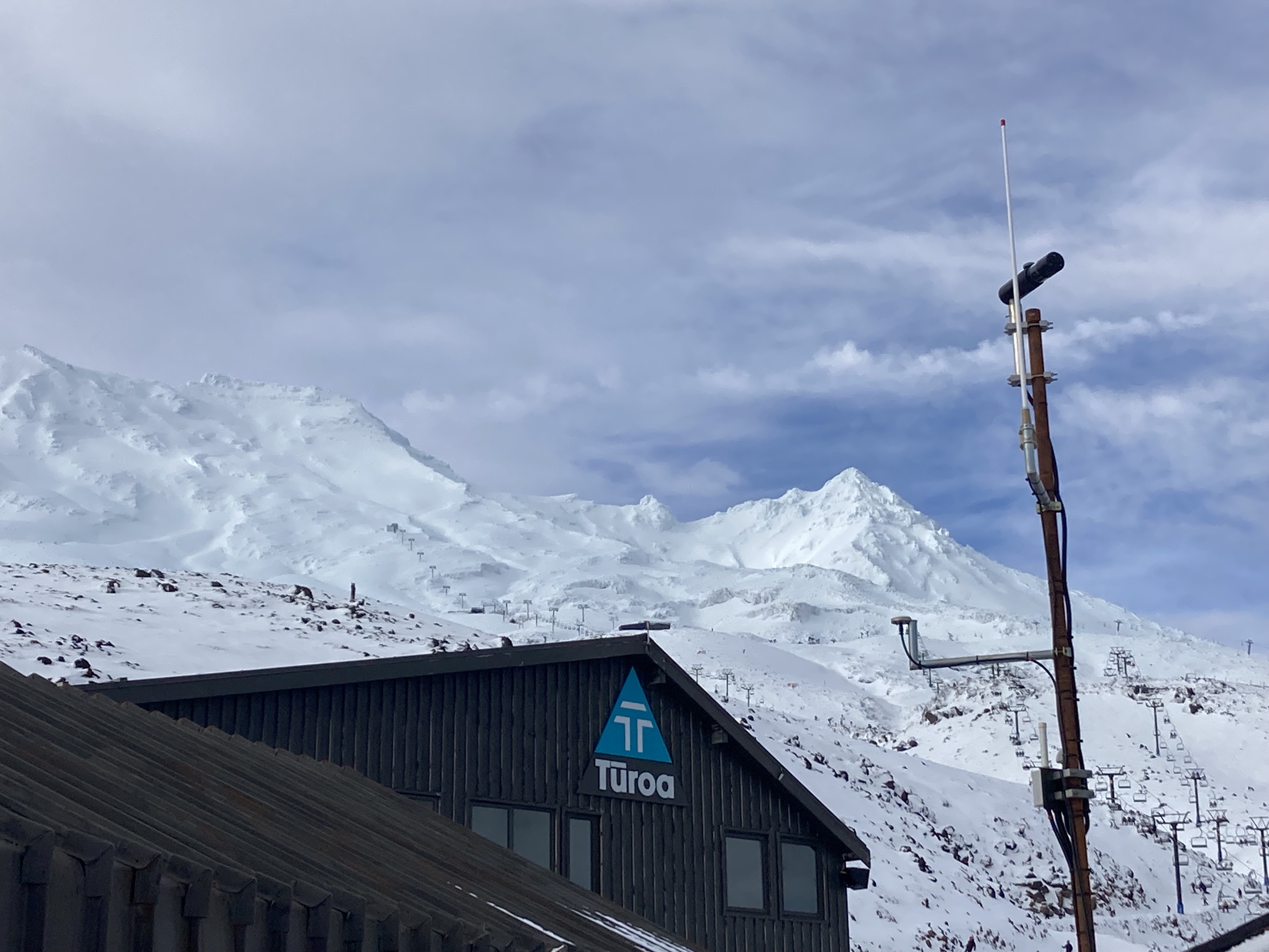 A scanDOAS scanner is mounted on the top of pole at a skifield building at Ruapehu. The skifield and the summit of Ruapehu are in the background.